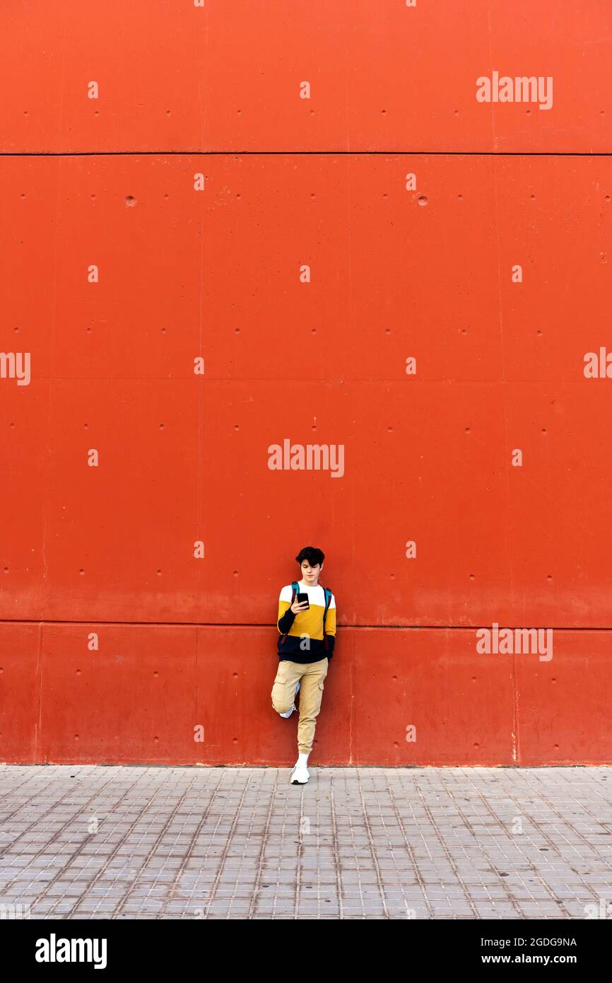 Young male with backpack standing on red wall using a mobile phone Stock Photo