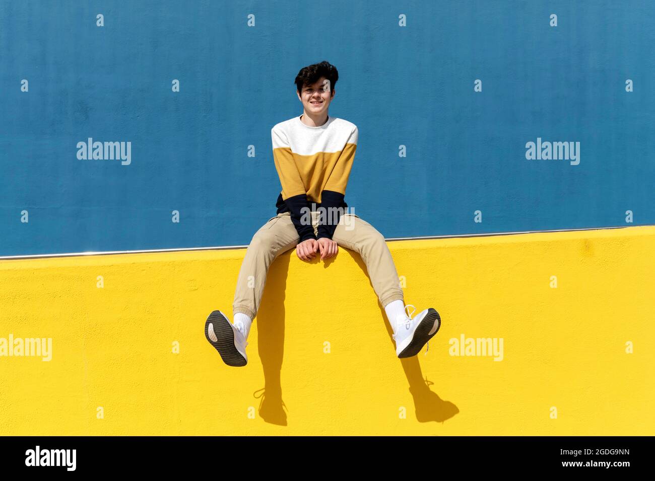 Confident man looking to camera while sitting on yellow retaining wall Stock Photo
