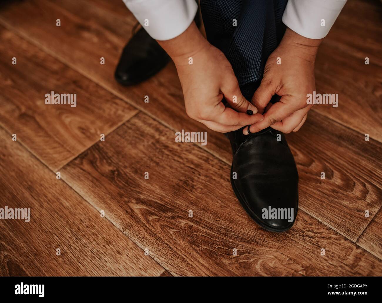 A man in a white shirt and dark trousers is sitting on a chair tying the laces of his black boots on a brown textured shiny wooden floor. Men's Stock Photo