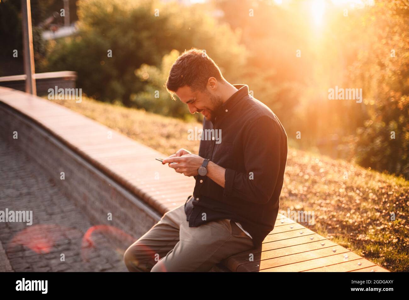 Happy man using smart phone sitting on bench at sunset Stock Photo