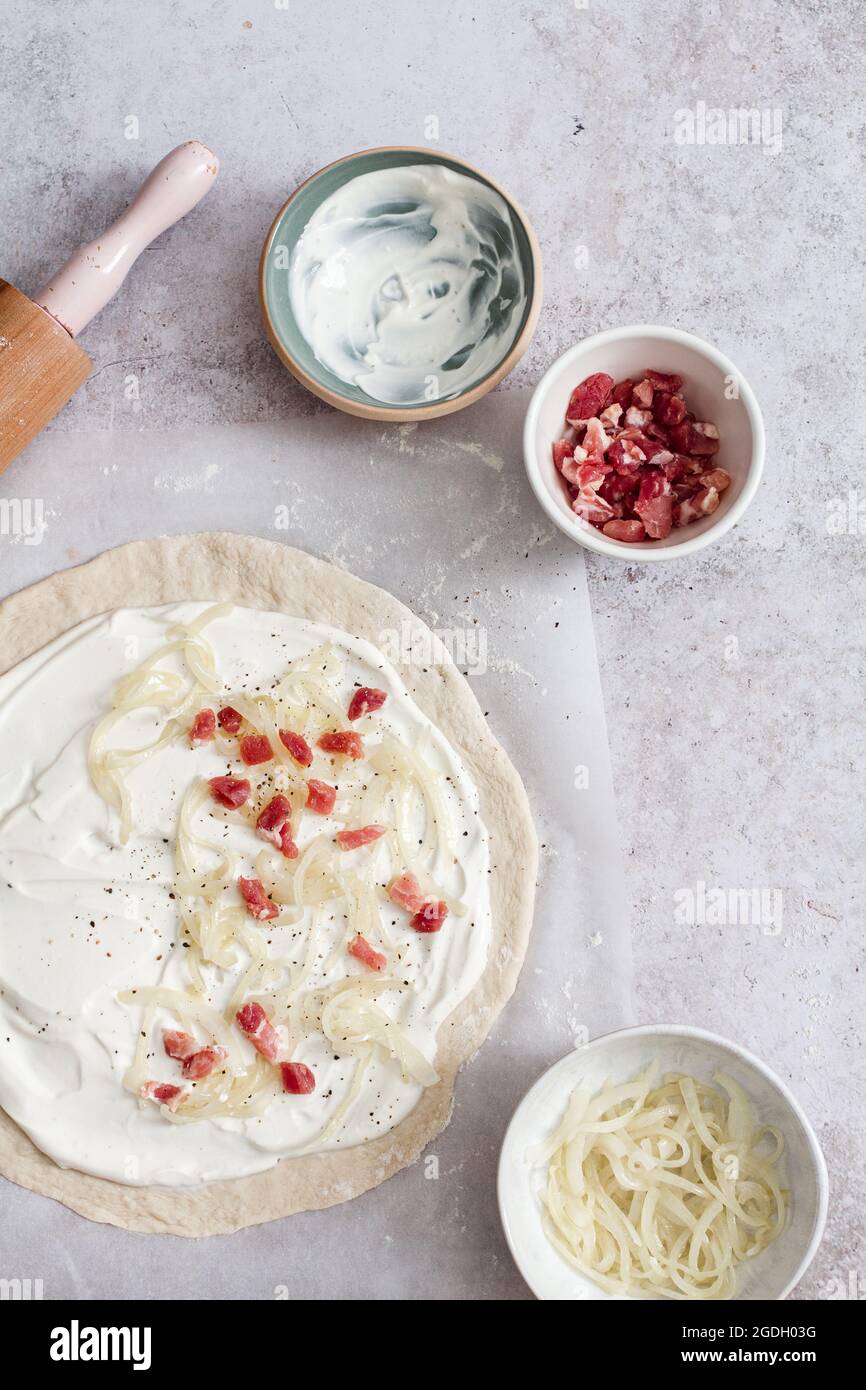 Preparation of a traditional tarte flambee (flammkuchen) with creme fraiche, fromage blanc, lardons and onion. Stock Photo