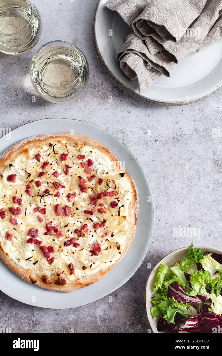 Tarte flambee (flammkuchen) with creme fraiche, lardons and onions served with a bowl of side salad and two glasses of white wine. Stock Photo
