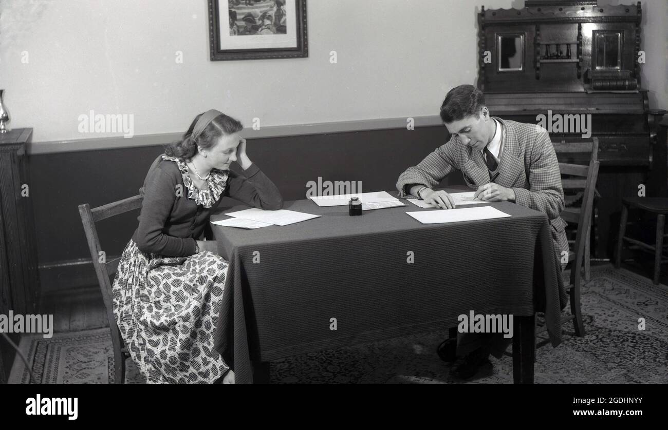 1950s, historical, inside a dinning room of the era, a couple sitting  on wooden chairs at the ends of a small table, covered with a tablecloth. Using a fountain pen and with an ink bottle on the table, the man iis writing on paper sheets, while the young lady is reading the handwritten manuscript Stock Photo