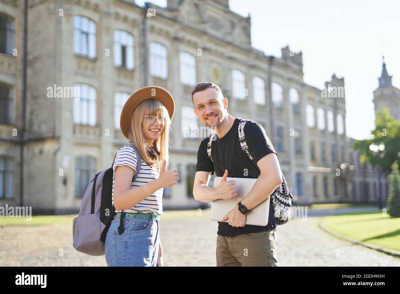 Two cheerful students, male and female, with backpacks raising thumbs up at the university campus holding laptop. Adorable successful students couple smiling showing thumbs up sign at the camera Stock Photo