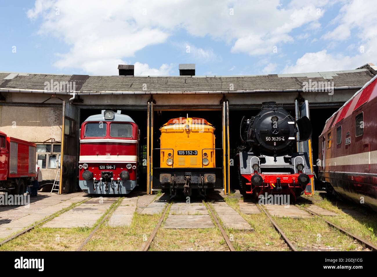 Electric railcars and a steam locomotive are standing in a locomotive shed in front of blue sky and clouds. Stock Photo