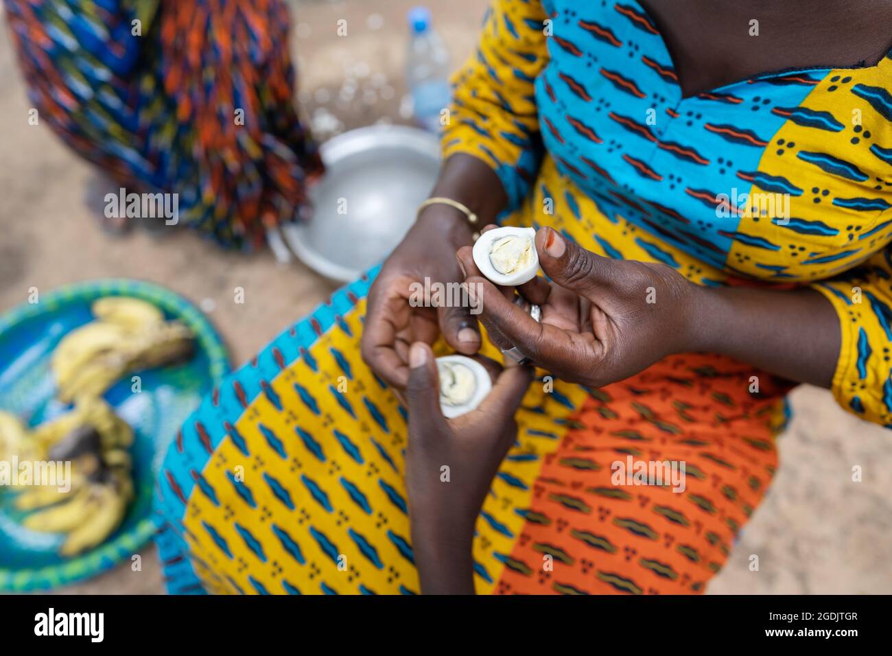Caring African woman in a colorful dress sitting on the floor in a rural village kitchen, sharing half of an egg with a child; poverty concept Stock Photo