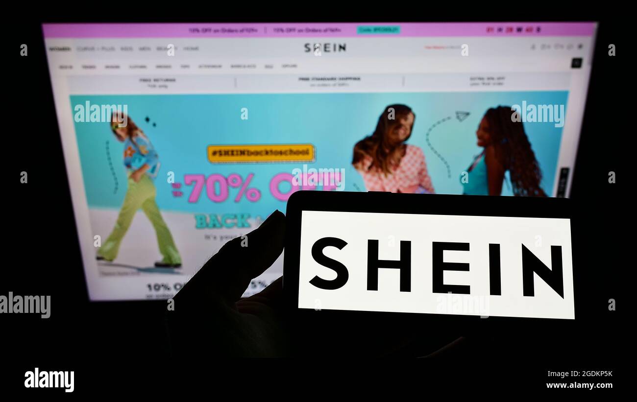 Person holding smartphone with logo of Chinese e-commerce company Shein on screen in front of website. Focus on phone display. Stock Photo