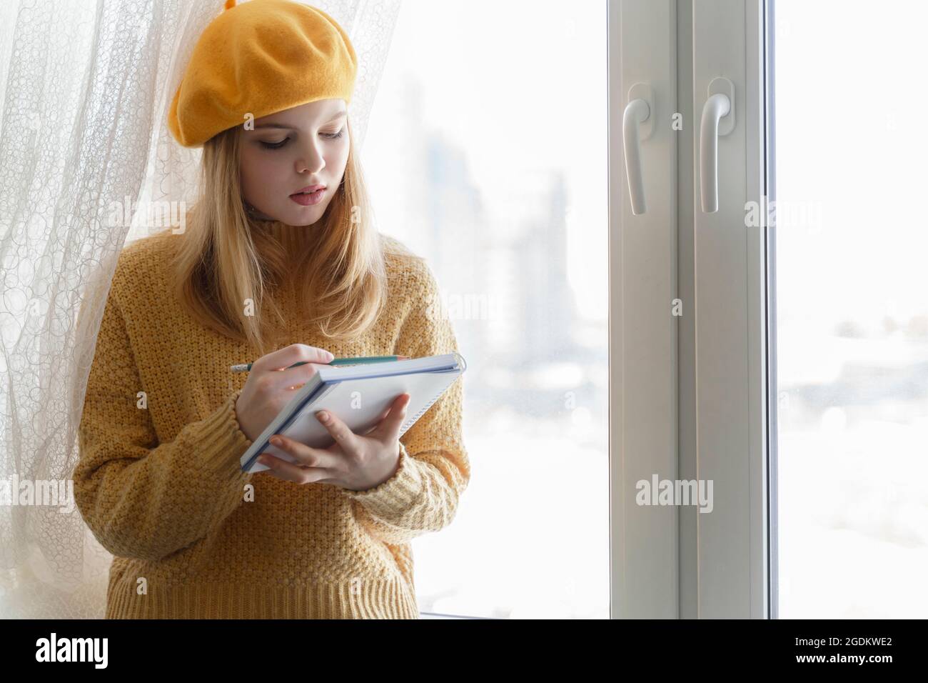 a young girl artist makes sketches in her sketchbook sitting by the window in a yellow beret Stock Photo