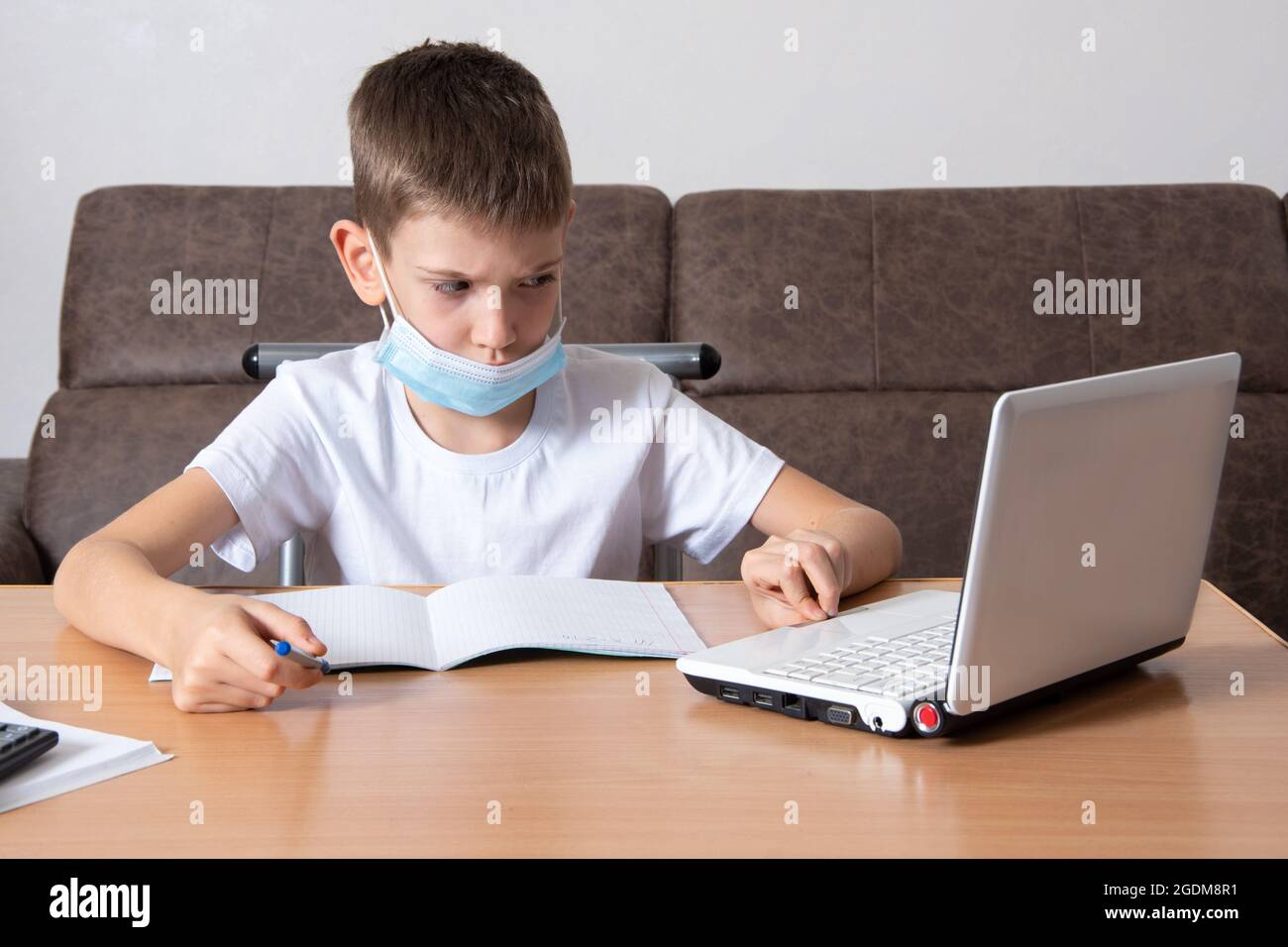 Distance learning during an epidemic - A serious schoolboy in a protective mask on his face looks at a laptop, writes information in a notebook, sitti Stock Photo