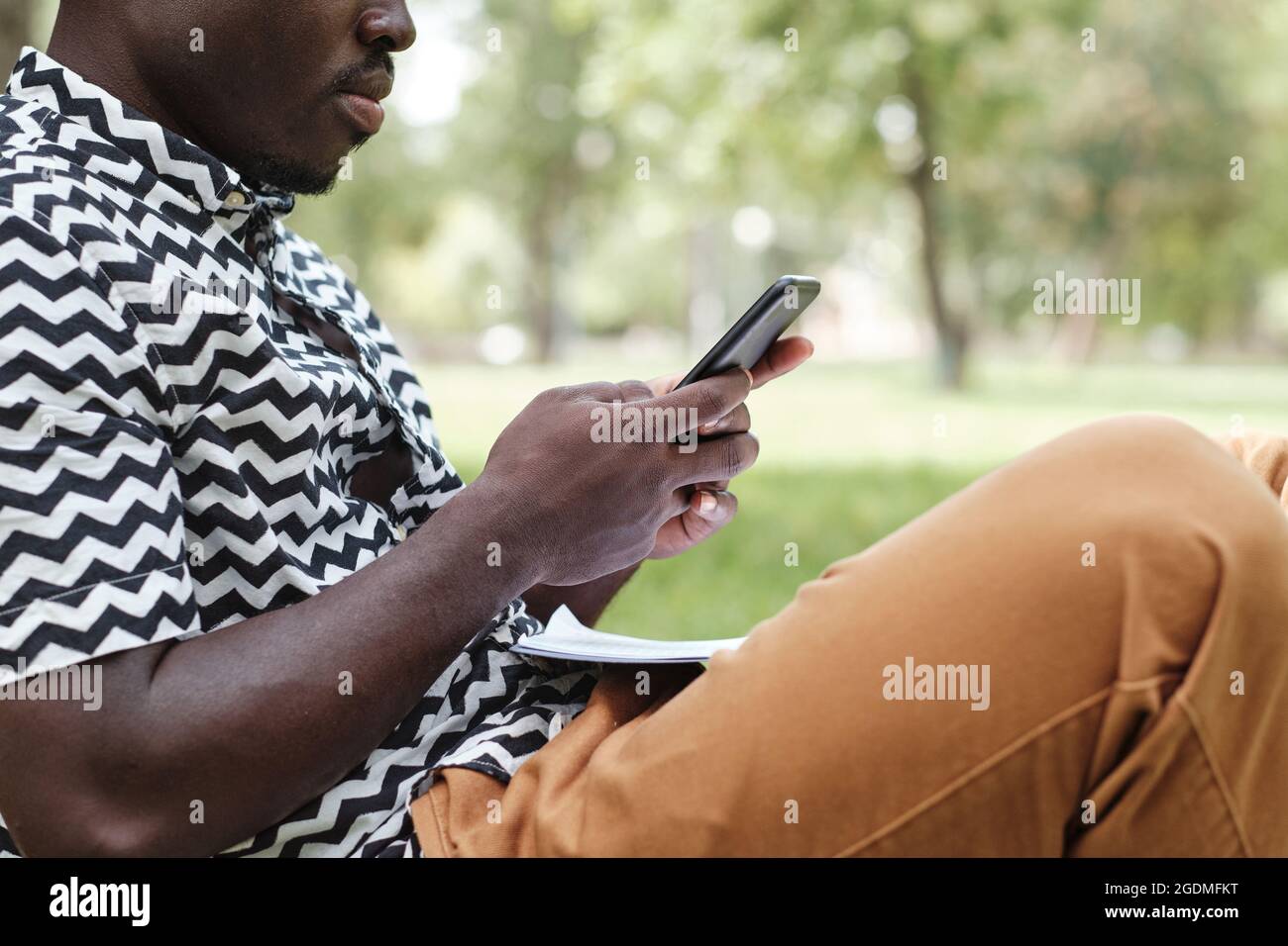 Close-up of African man typing a message on his mobile phone while sitting in the park outdoors Stock Photo