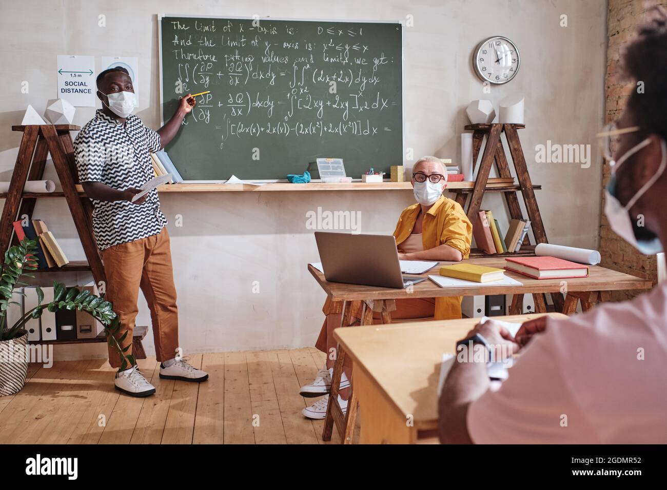 African student in mask standing near the blackboard and pointing at it ...