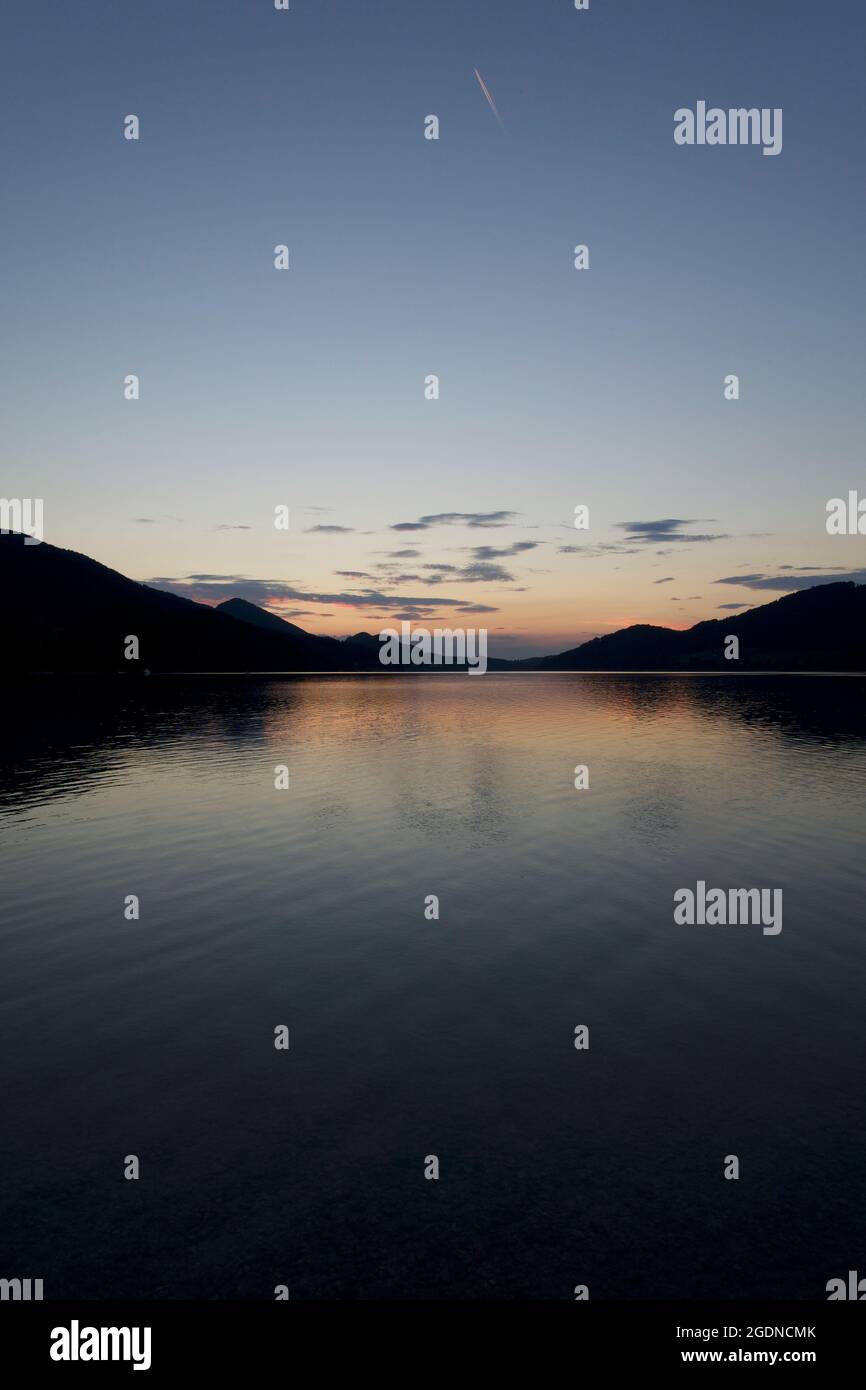 Shooting star saying good night to the orange sunset reflected at the foot of the Alpine mountain range in the calm and translucent lake water. Stock Photo