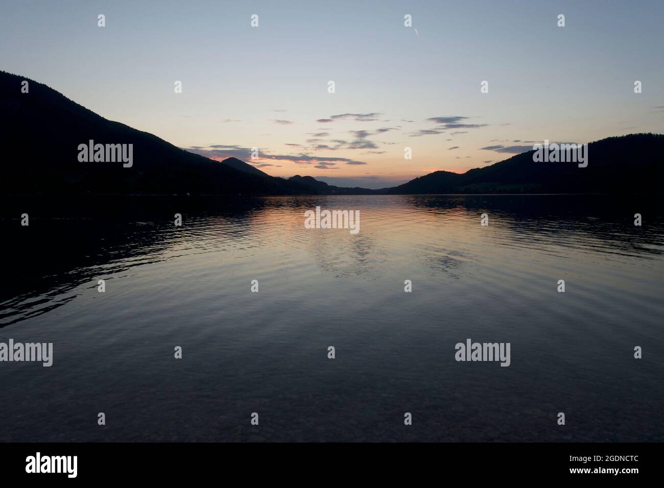 Clear lake water showing through the pebbles and rippling out towards the last sunset reflections of day's end at the foot of the Alpine mountains. Stock Photo