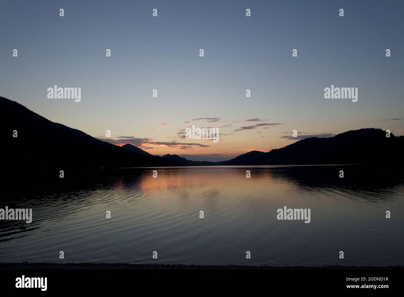 Atmospheric centered waterscape with the last vestiges of the setting sun reflected in the rippled clear lake water surrounded by Alpine mountains. Stock Photo