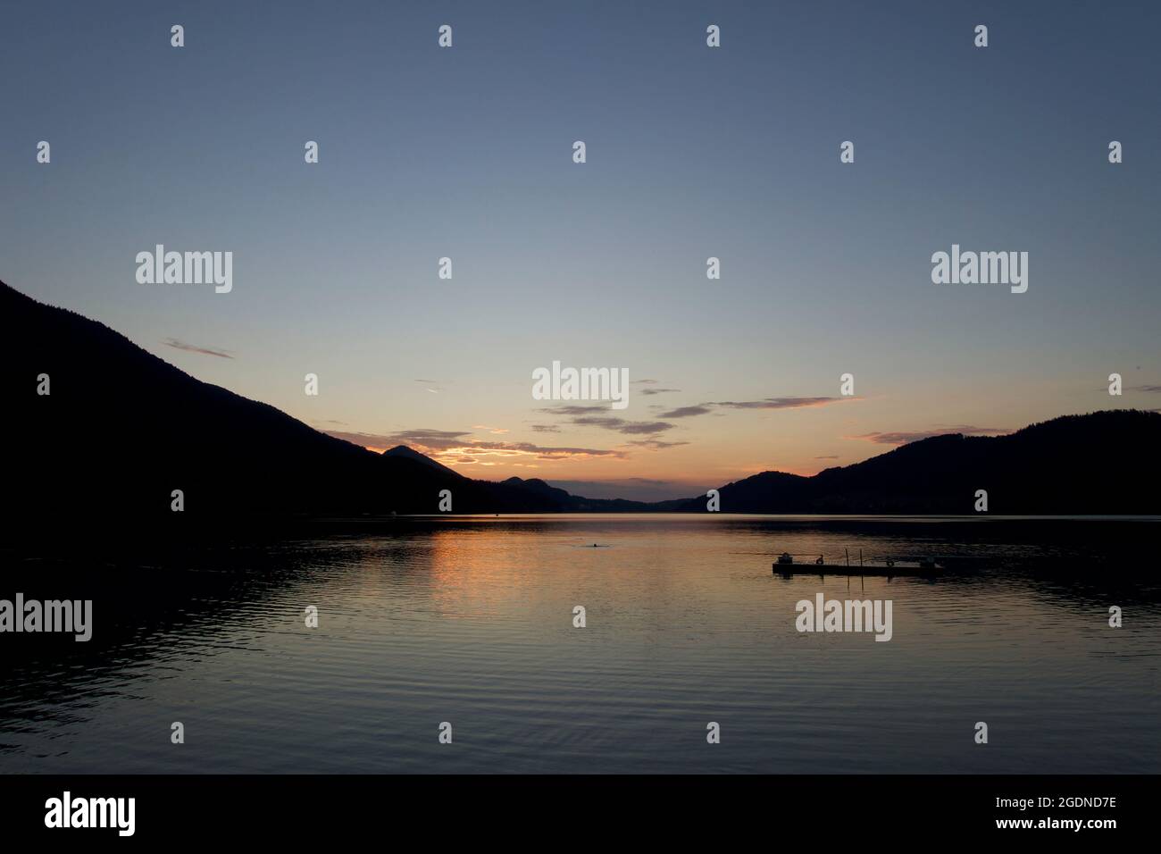 Almost day's end. Dark blue sky, orange setting sun on the horizon sandwiched between two silhouetted Alpine mountain ranges reflected in lake water Stock Photo