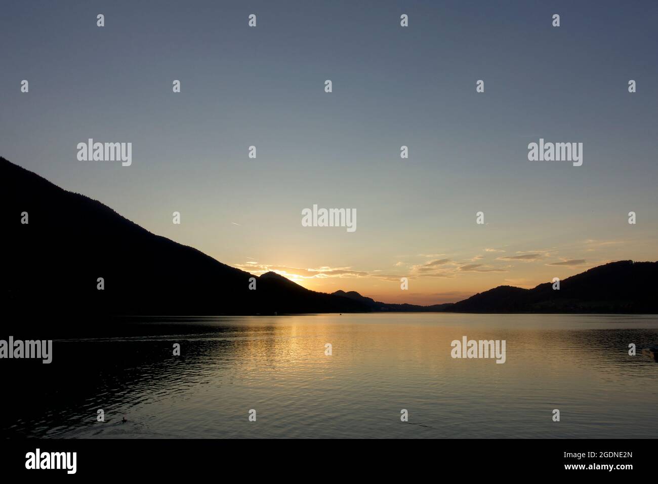 Picturesque late summer sunset. Low yellow clouds, orange coloured water ripples and lake reflections overlooked by Alpine Austrian mountain range. Stock Photo