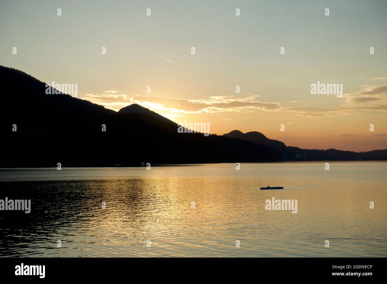 Silhouetted Alpine mountain range behind tranquil lake with yellow clouds & setting sun. Blissful scenic spot with paddle board & buoy in the water Stock Photo