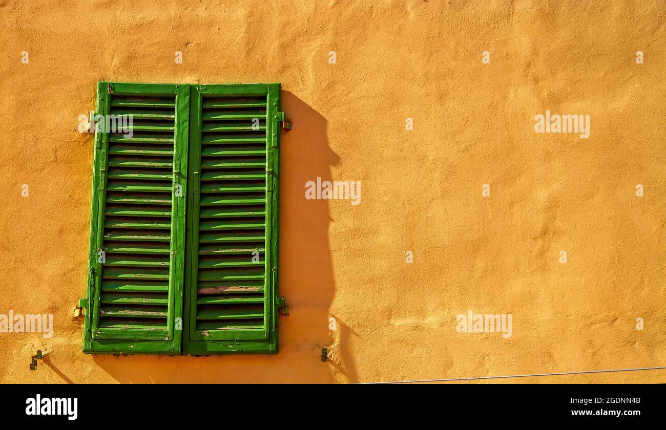 A pair of old, green, wooden shutters mounted on a terracotta painted building, and lit by some sunlight Stock Photo