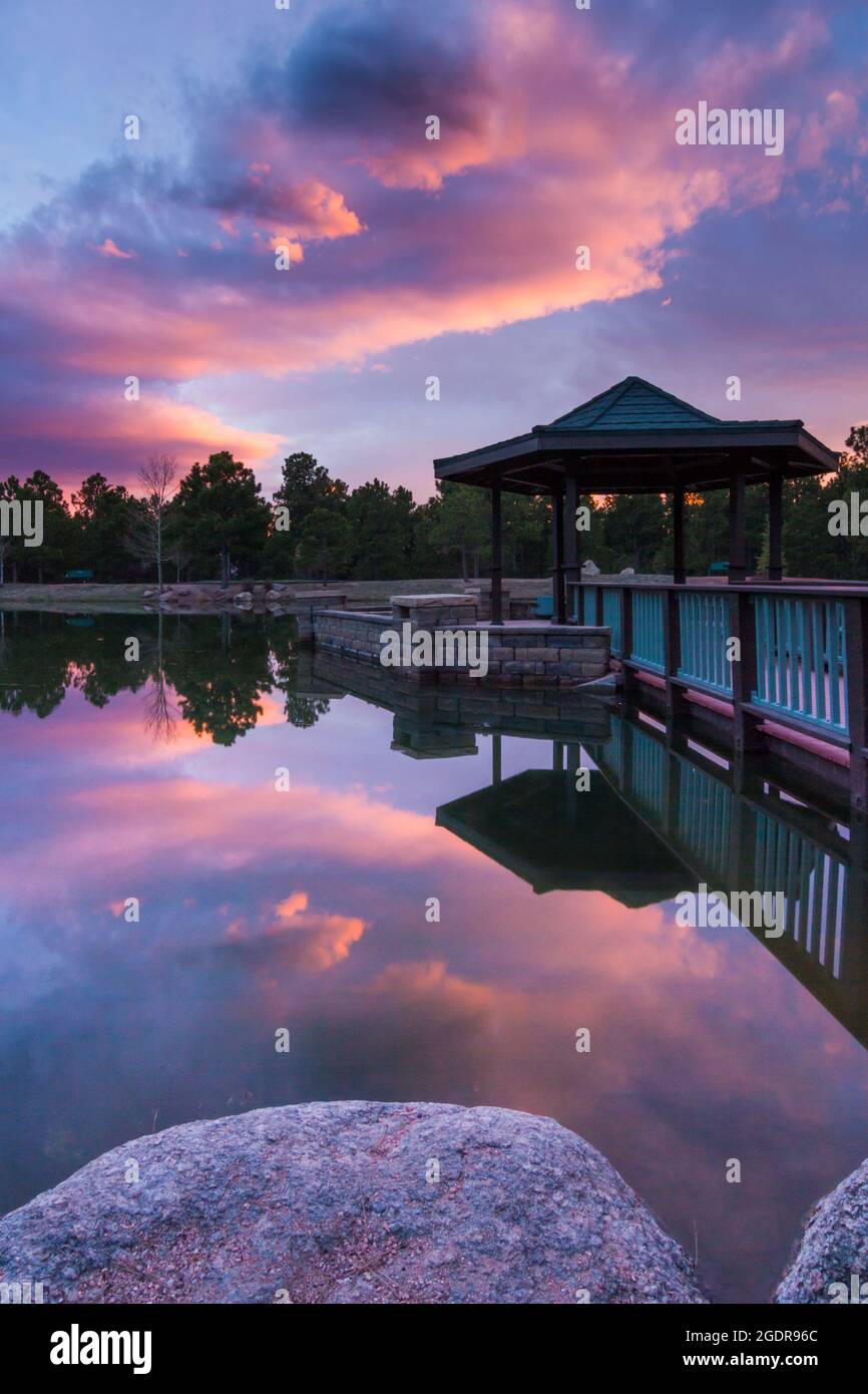 Glassy reflection of a brilliant pink and purple sunset over a gazebo in the middle of a pond in Fox Run Regional Park in central Colorado Stock Photo