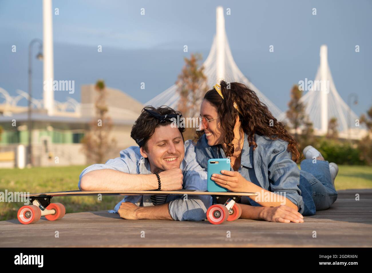 Happy smiling woman show man message on smartphone lying on skateboard outdoor in summer urban park Stock Photo
