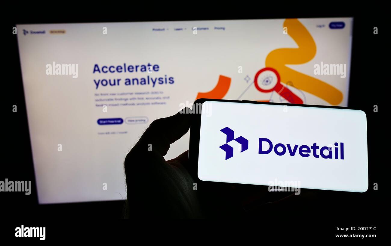 Person holding mobile phone with logo of customer research company Dovetail Research Pty. Ltd. on screen with web page. Focus on phone display. Stock Photo