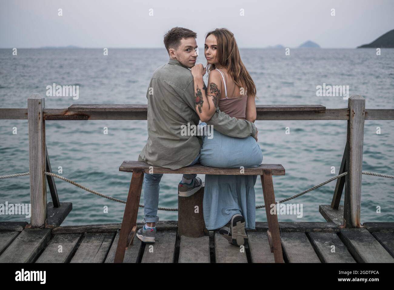 Beautiful young couple in love sitting on the bench by the sea, cuddling and looking at camera with copy space. High quality photo Stock Photo