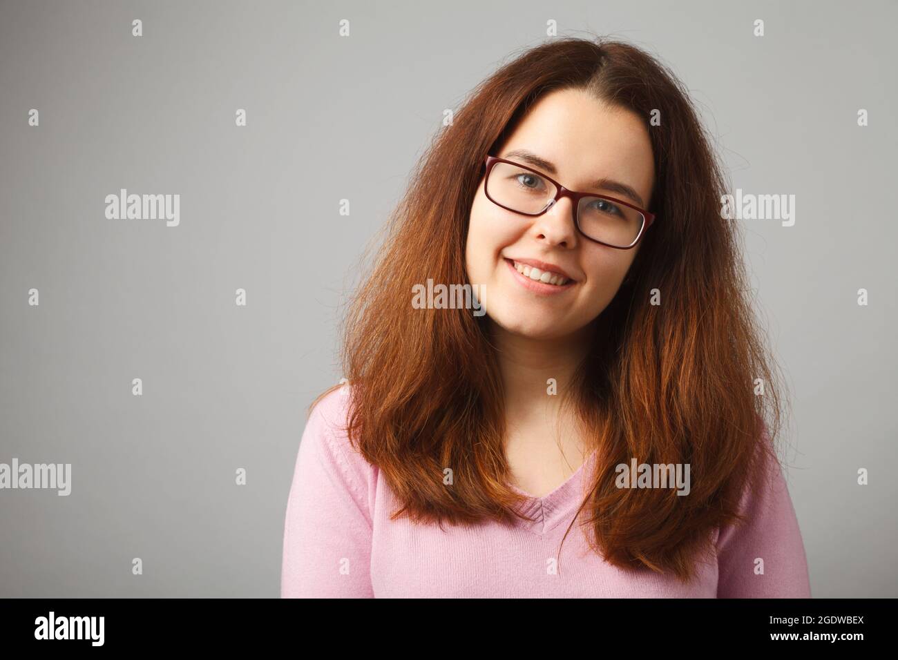 Redhead long-haired girl with glasses smiles, looking at the camera. Studio portrait of a girl on a solid light background. Stock Photo