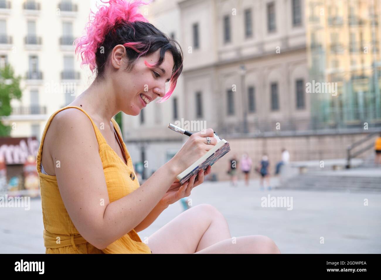Young caucasian woman with pink hair sitting outdoors writing on a ...