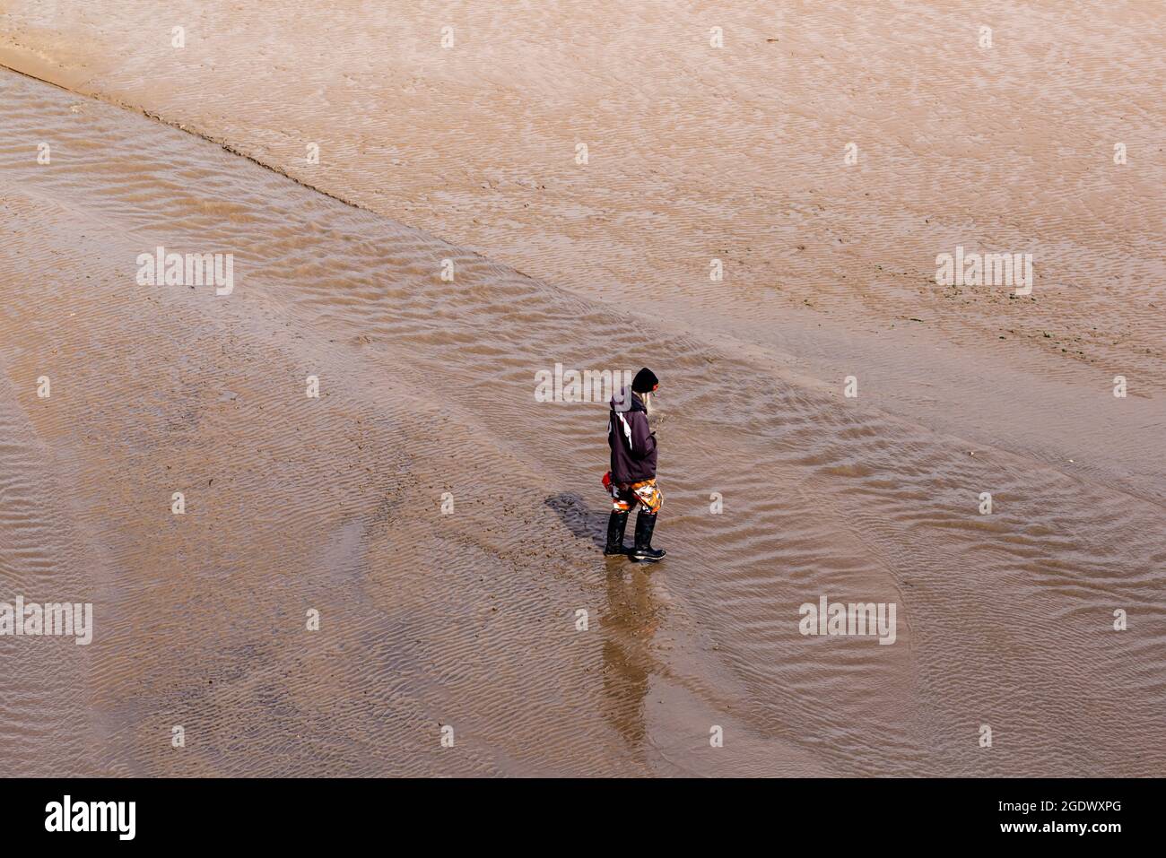 Woman in hat and wellingtons looking down walking on a sandy beach through a stream of water Stock Photo