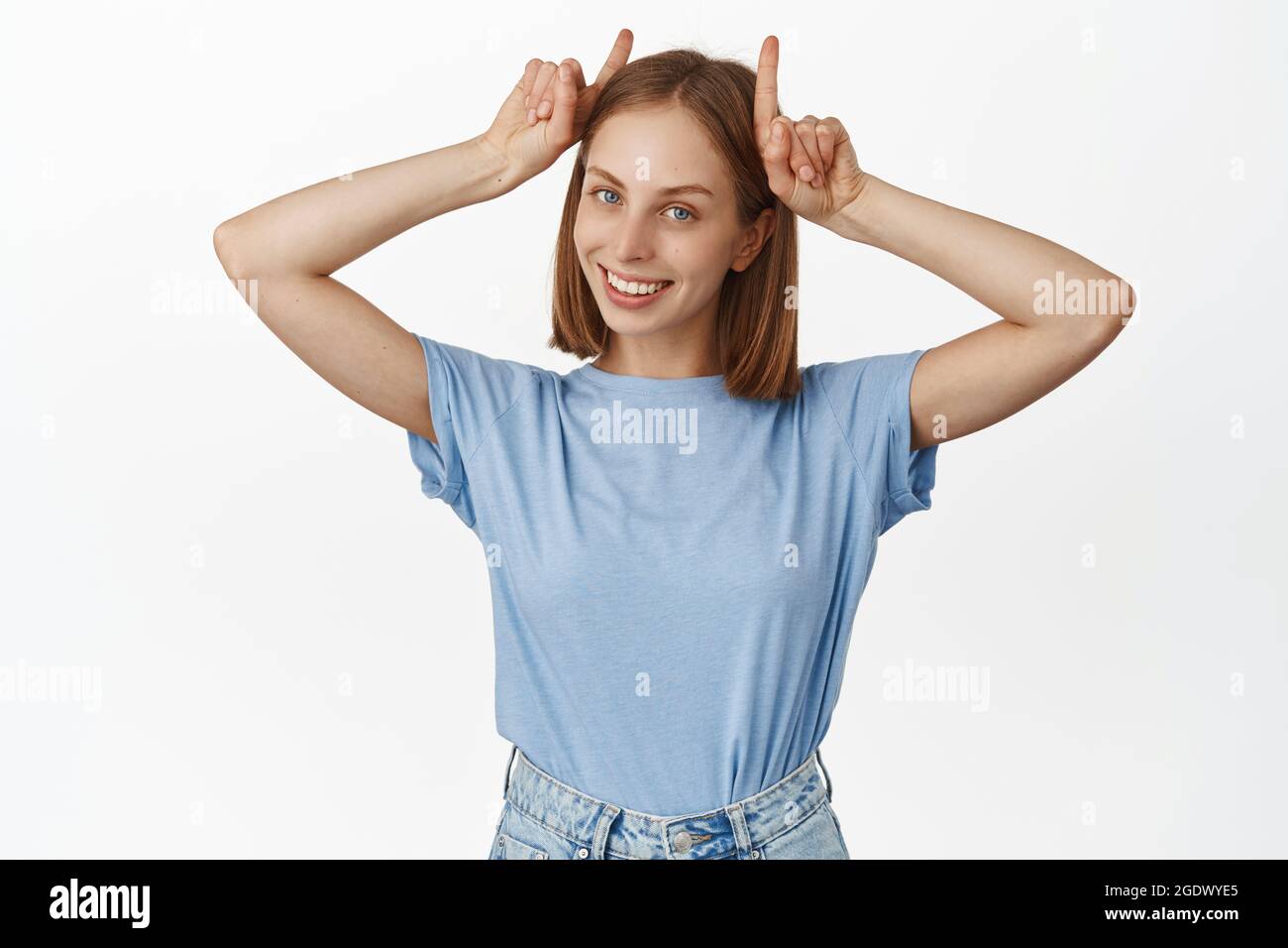 Image of silly and cute young woman having fun, showing bull horns gesture on head and smiling happy, being playful, standing in t-shirt against white Stock Photo