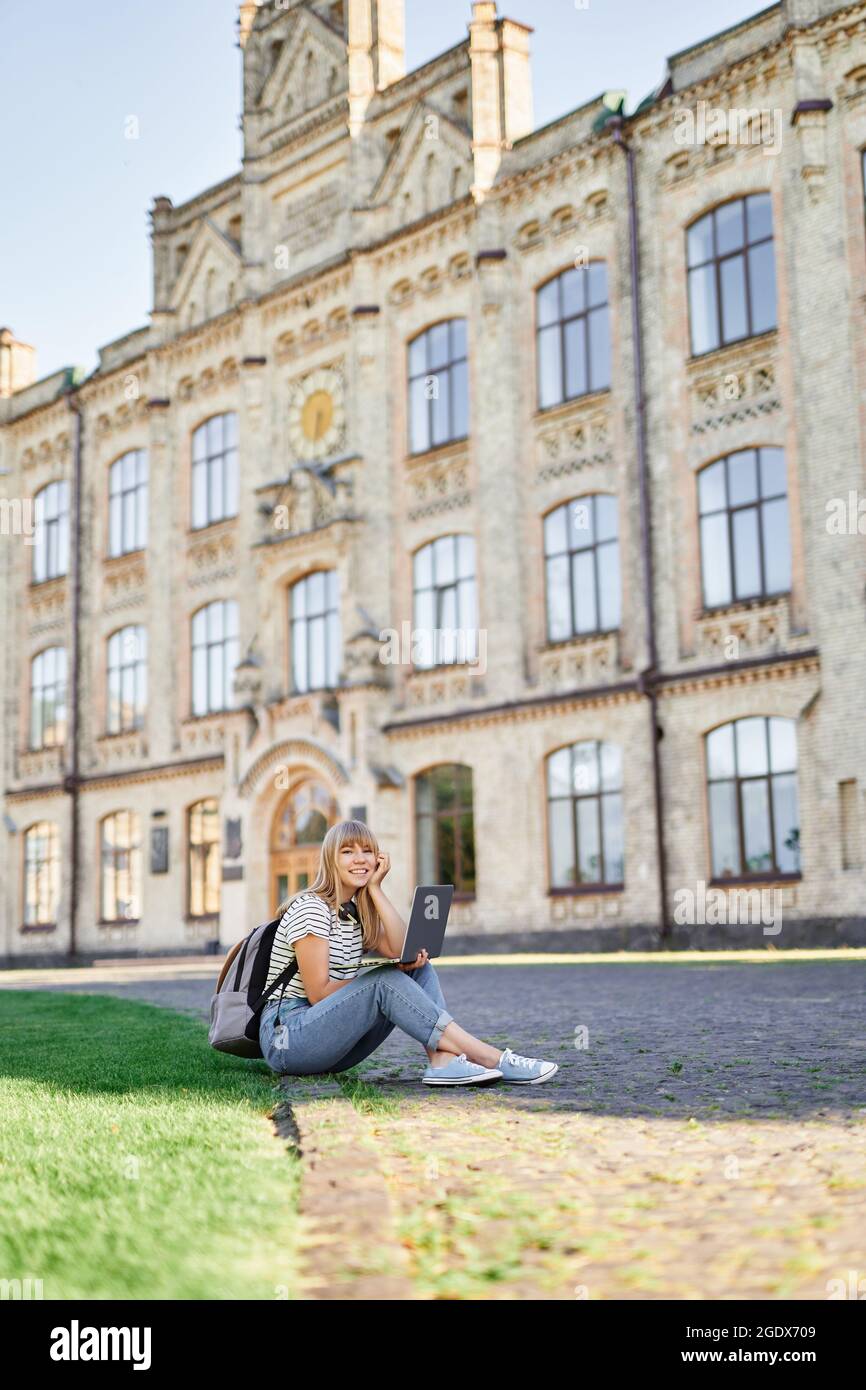 Happy school girl, e-learning, study online, student portrait or education concept. Cheerful young blonde teen university or college student with backpack and laptop sitting on green grass in campus.  Stock Photo