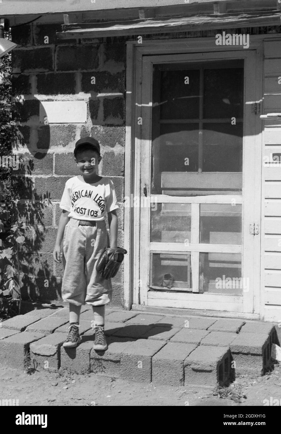 1950s, historical, starting young, it's summertime and outside the door to his home, a small boy standing for a photo in his basebal kit, cap and with pitchers glove, Des Monies, USA. On his baseball shirt is the wording 'American Legion'. Founded in 1925, American Legion Baseball was a program set up to provide a national baseball tournament for teenagers, with the objective to teach young Americans the importance of sportsmanship, good health and active citizenship. Stock Photo