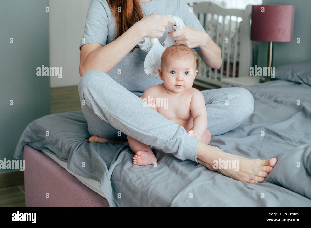 A young mother is sitting on the bed and dressing up her infant baby