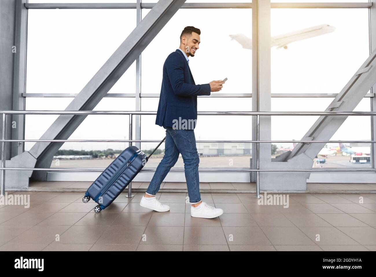 Handsome arab guy using smartphone while walking with suitcase at airport terminal Stock Photo