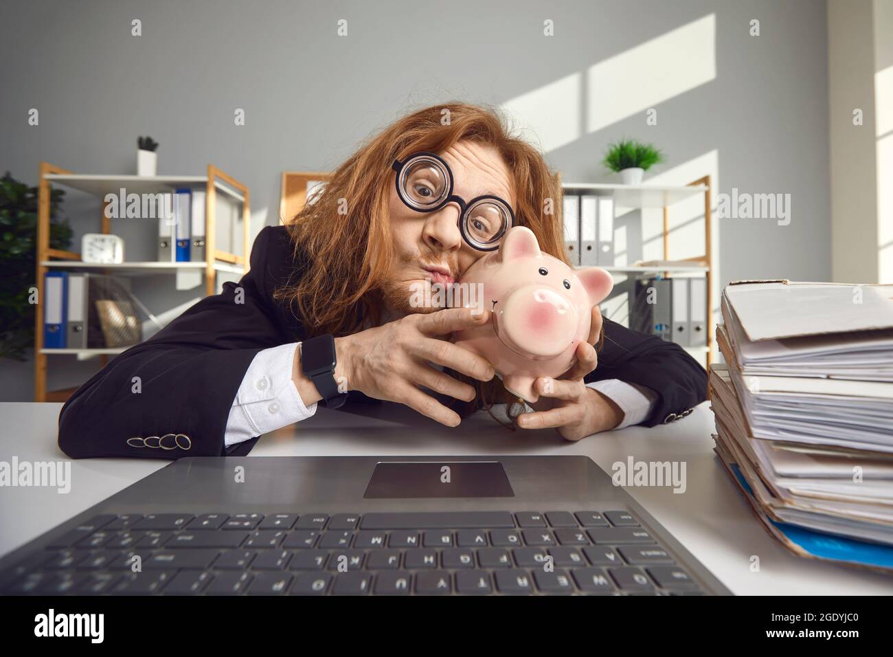Strange freak guy in big round glasses kisses a piggy bank while sitting at office desk with laptop. Stock Photo