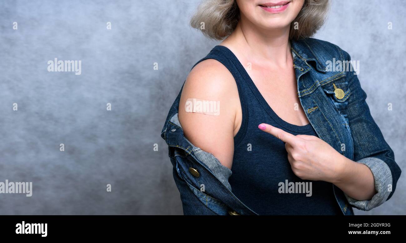 Woman showing arm with plaster after getting COVID-19 vaccine. Coronavirus vaccinated healthy person points shoulder on gray background. People vaccin Stock Photo