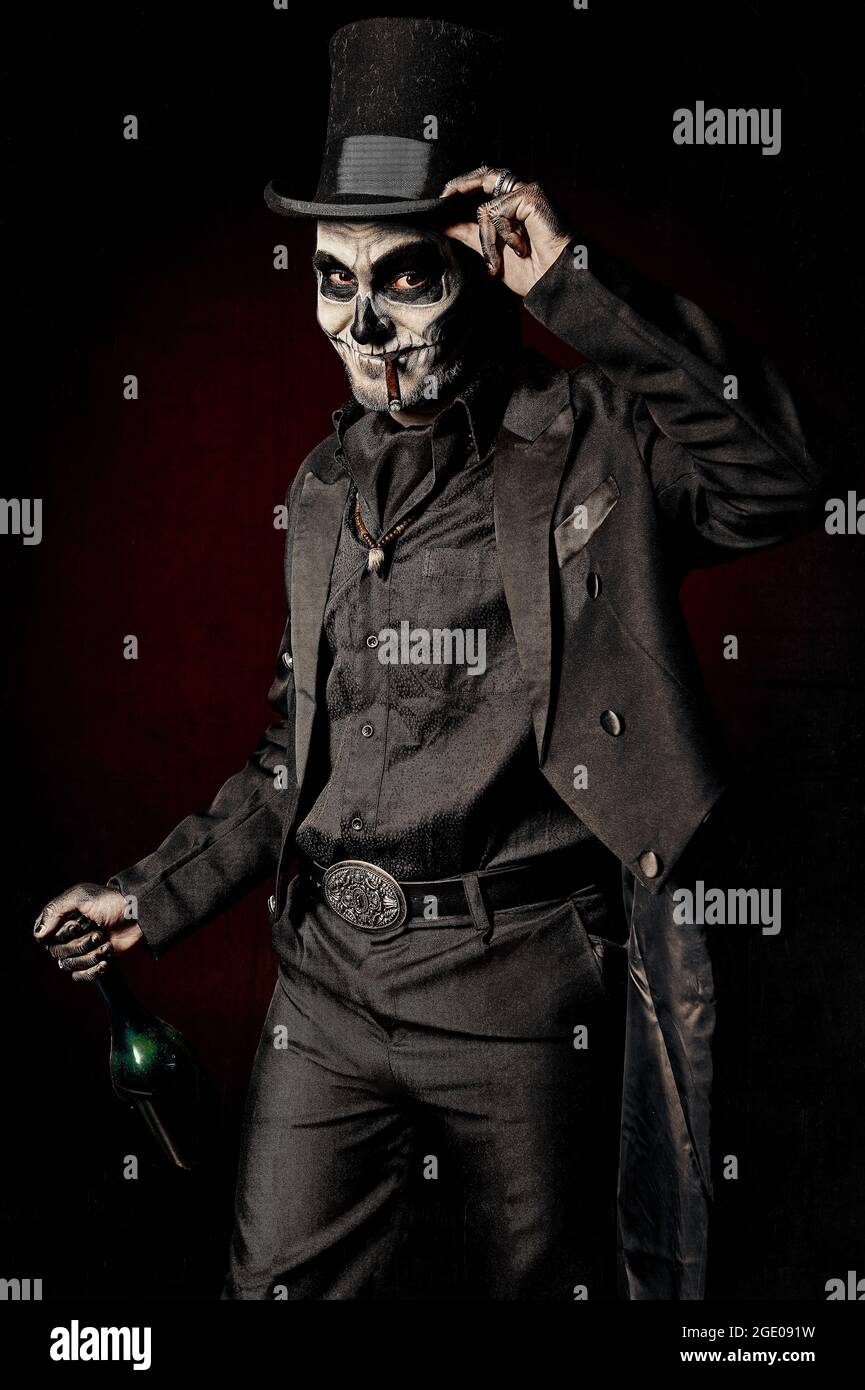 A young man in image of Baron Samedi, the Voodoo deity. Baron Saturday in black suit and top hat with a bottle of alcohol in hand. Day of the Dead (an Stock Photo