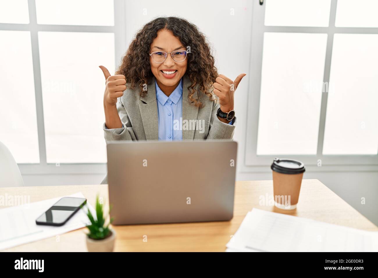 Beautiful hispanic business woman sitting on desk at office working ...