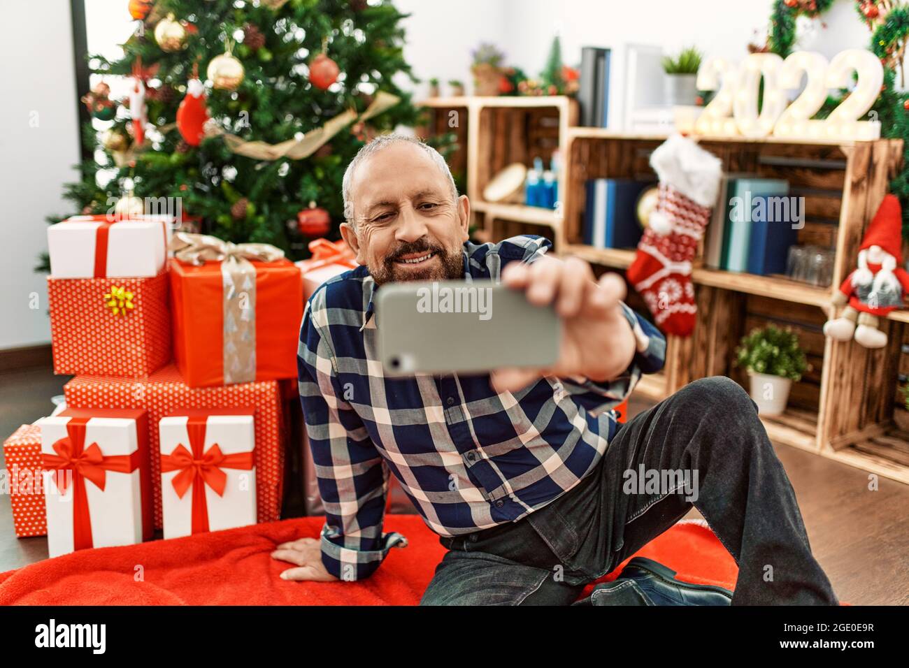 Senior man with beard wearing santa claus hat celebrating christmas at home, sitting on the floor by christmas tree and presents taking a selfie pictu Stock Photo