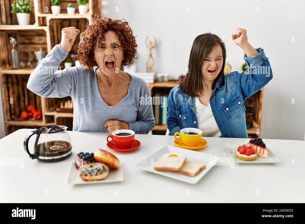 Family of mother and down syndrome daughter sitting at home eating breakfast angry and mad raising fist frustrated and furious while shouting with ang Stock Photo