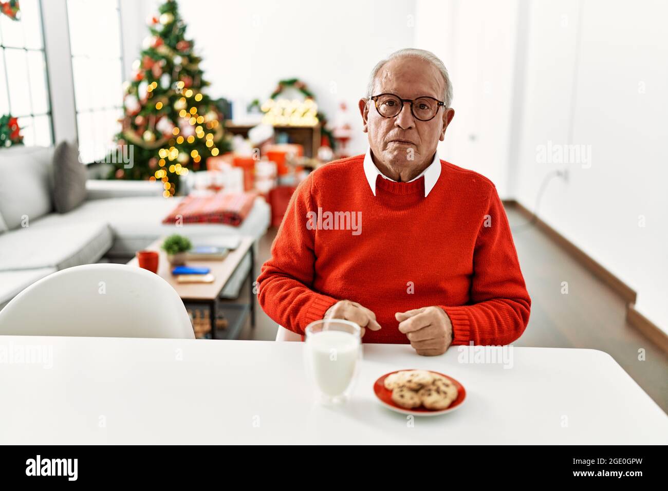 Senior man with grey hair sitting on the table with cookies by christmas tree with serious expression on face. simple and natural looking at the camer Stock Photo