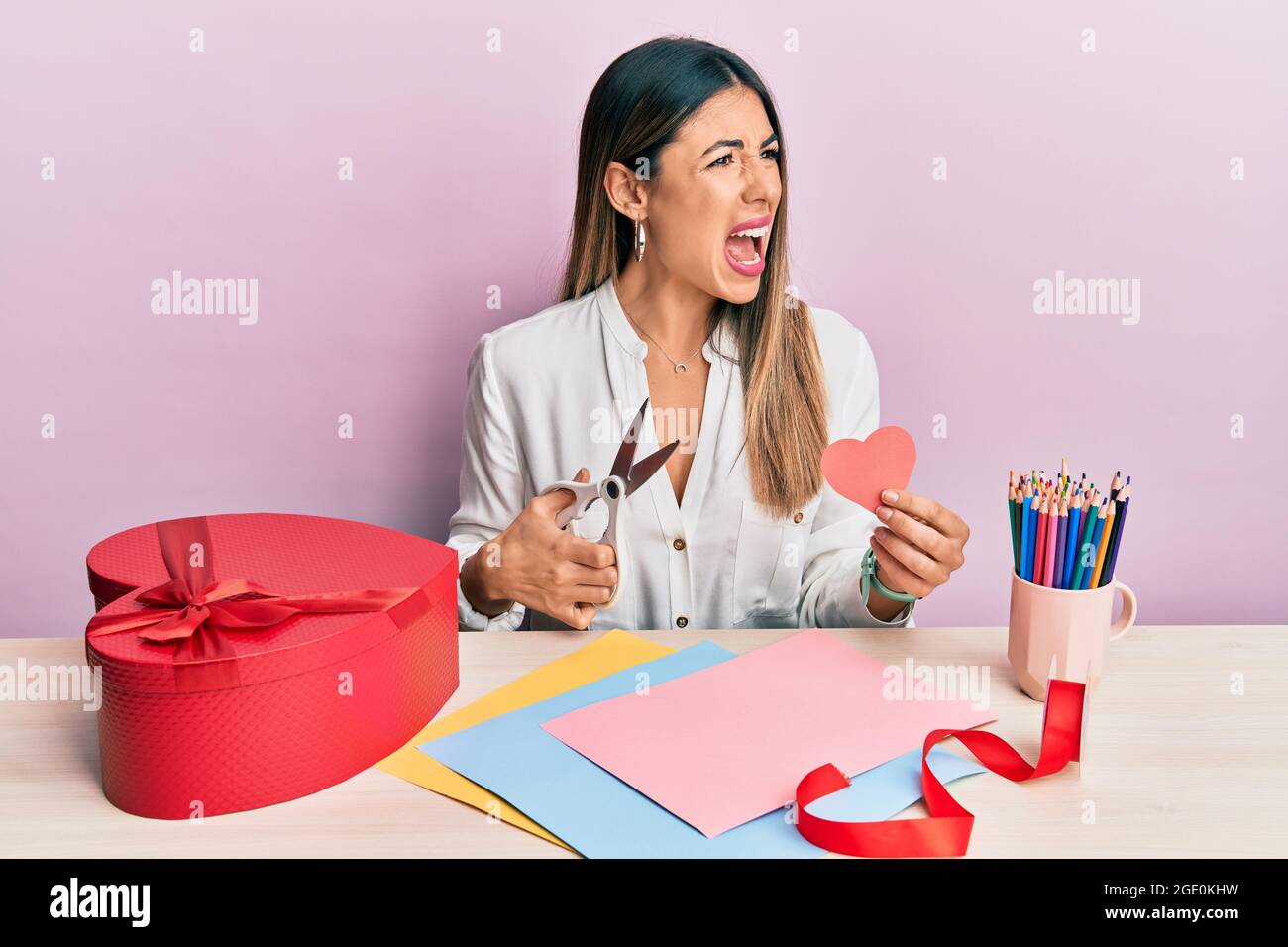 Young hispanic woman making valentine gift sitting on the table angry ...