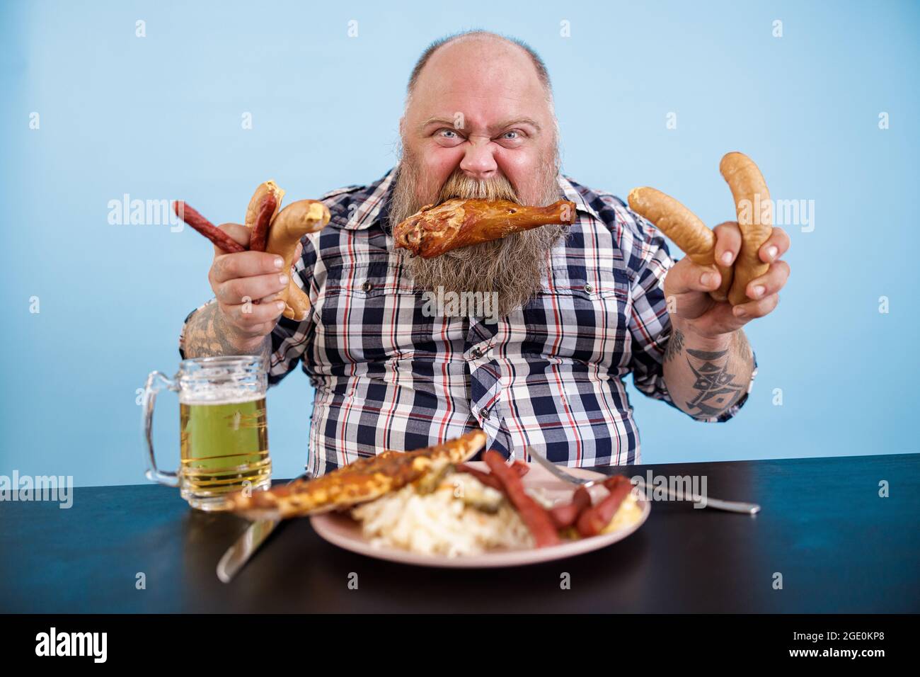 Fat man with chicken leg in mouth holds sausages at table in studio Stock Photo