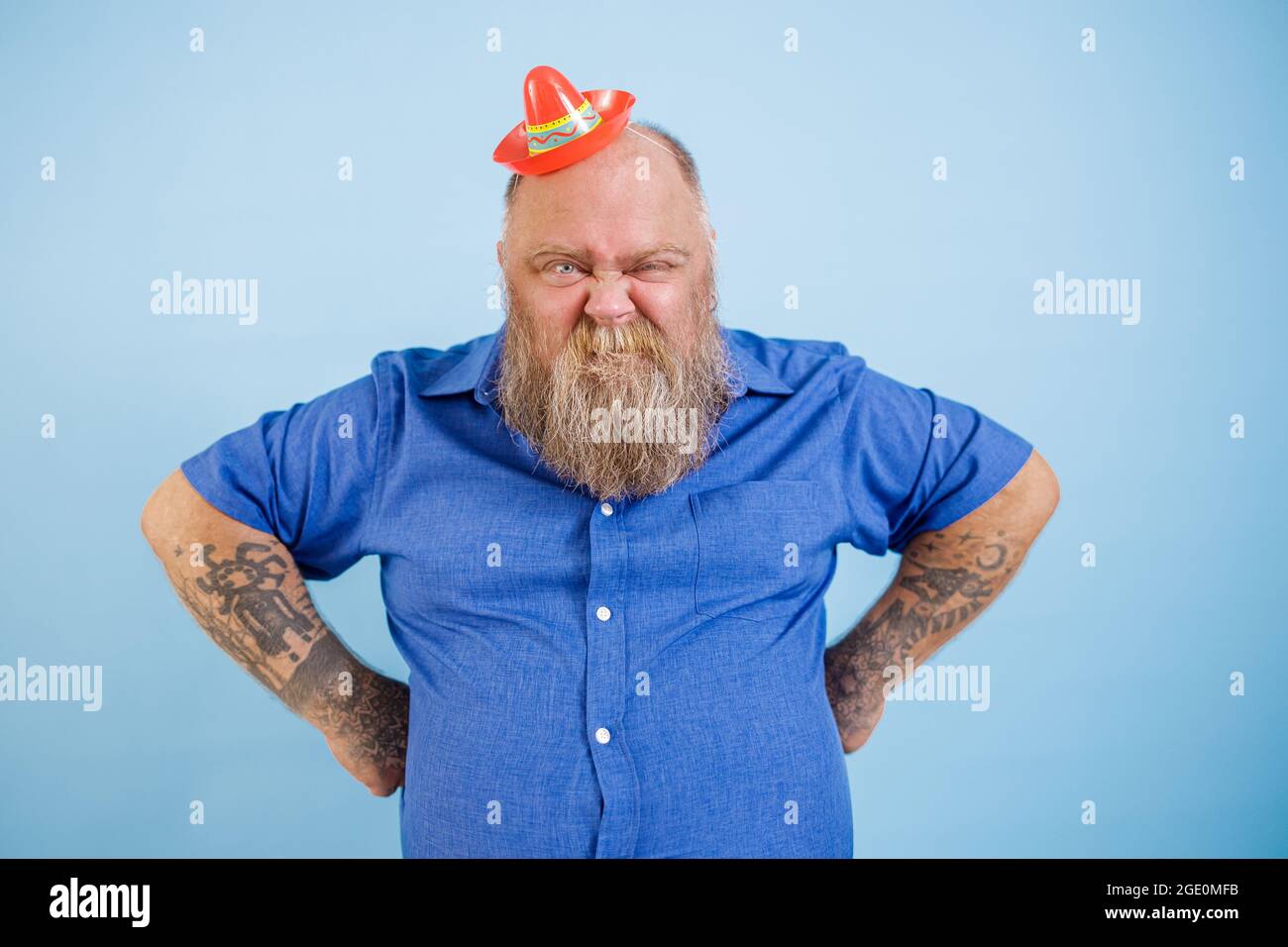 Funny plus size man wearing small sombrero holds hands on waist grimacing on blue background Stock Photo