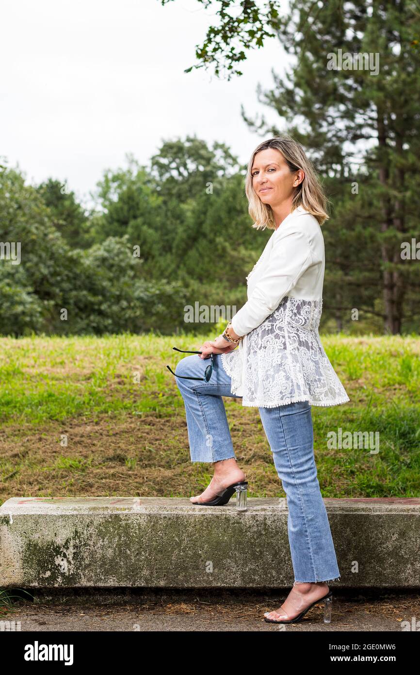 Full length portrait of a blonde haired model posing standing on a stone bench in a park in Spain.The woman is wearing jeans, a white jacket and trans Stock Photo