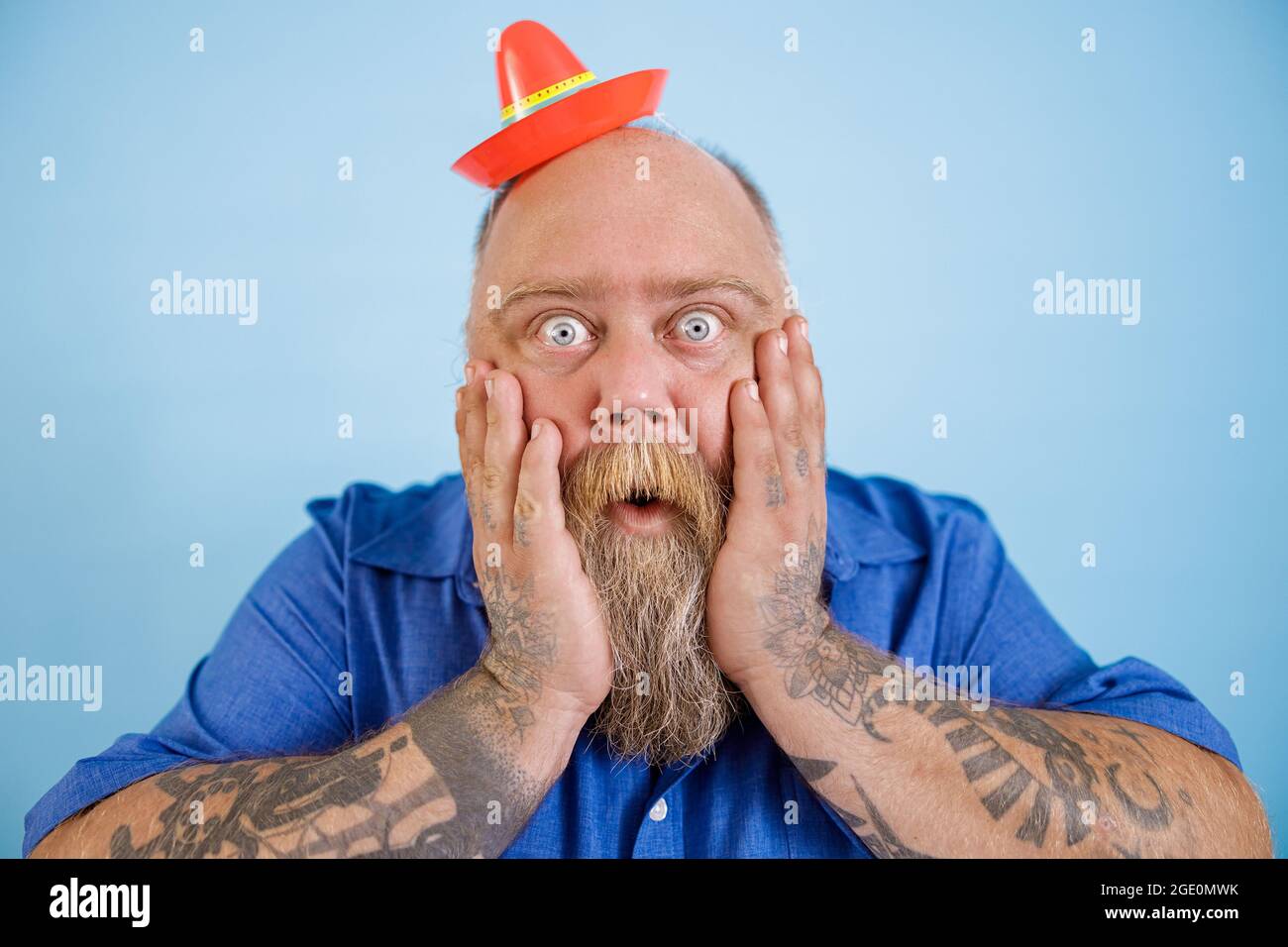 Shocked obese man with small sombrero holds cheeks on light blue background Stock Photo