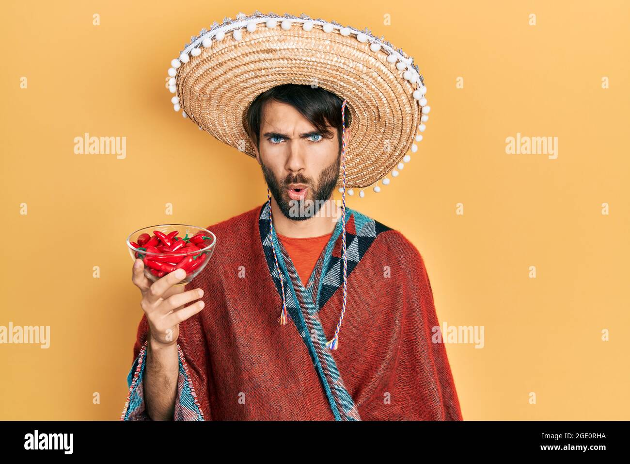 Young hispanic man wearing mexican hat holding chili scared and amazed with open mouth for surprise, disbelief face Stock Photo