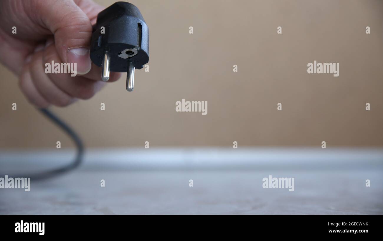 A 220 volt hybrid CEE 7/7 electric plug made of black plastic held by a man's hand right above the floor in low angle view image with copy space Stock Photo