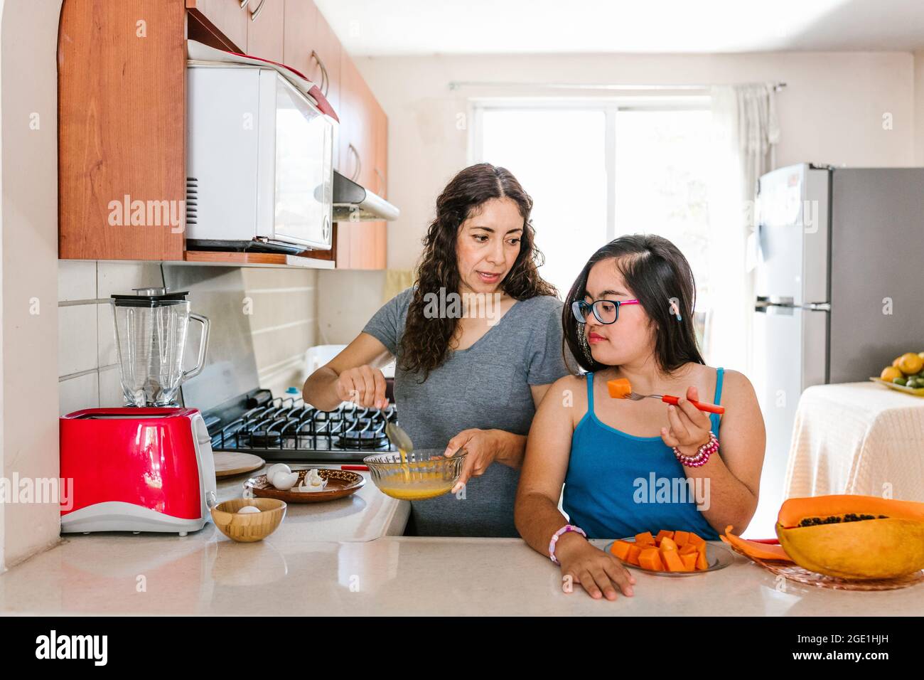 Mexican teen girl with down syndrome eating fruit and her mother cooking at home, in disability concept in Latin America Stock Photo
