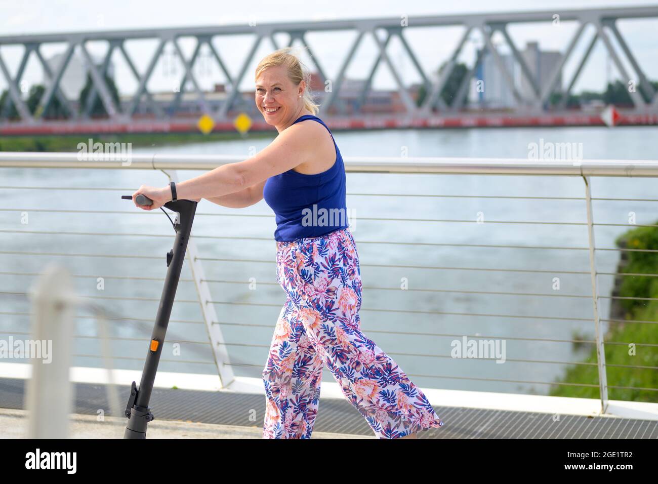 Happy vivacious middle-aged blond woman on an electric scooter crossing a bridge over a river on a hot summer day in an urban mobility or transportati Stock Photo